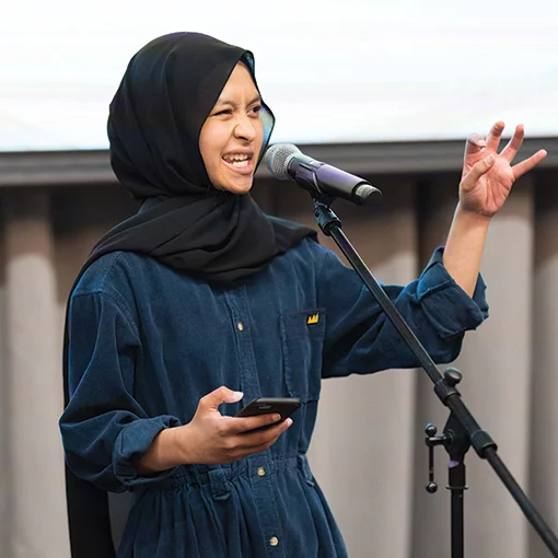A young woman wearing a black hijab and dark blue boiler suit speaking into a microphone.