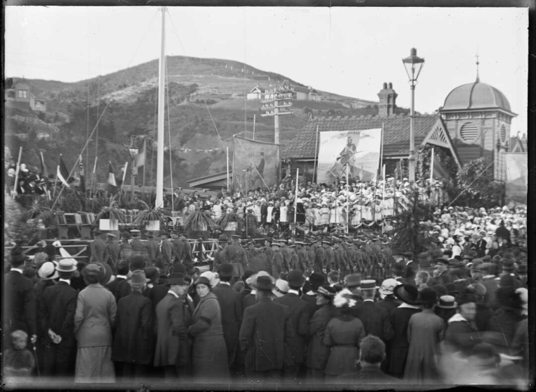 Shows a crowd, flagpole and banners next to the Petone Railway Station.