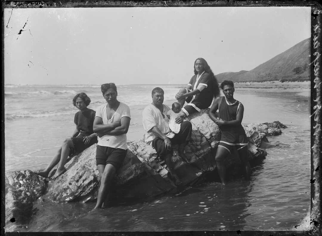 Shows a family including small baby sitting atop rocks at the shore, some members are standing in the water.