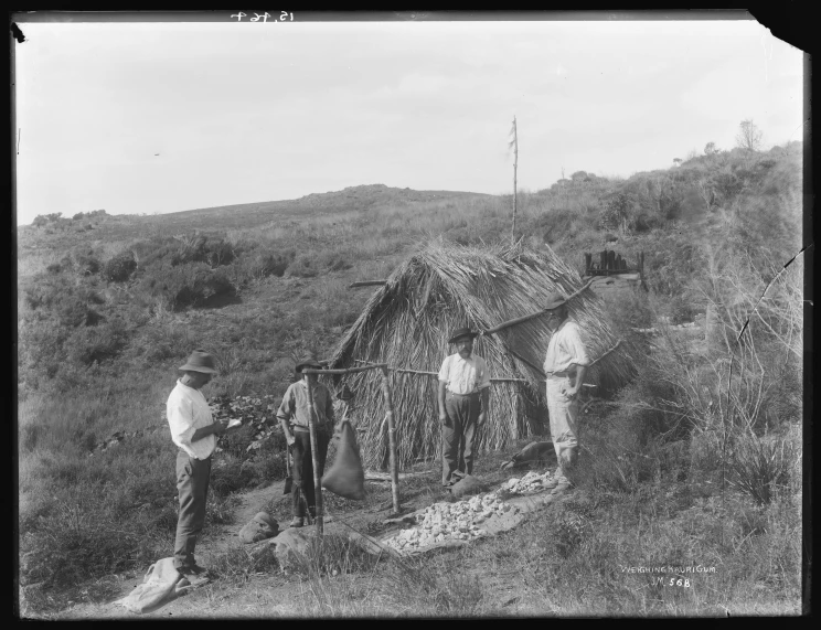 A group of men are seen weighing gum with a small thatched hut in the background.