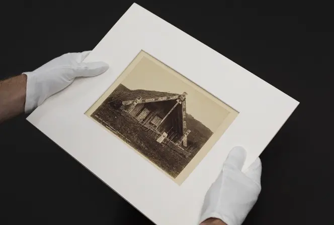 Gloved hands holding a photo of the whare belonging to King Tawhiao at Te Kuiti.