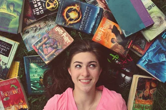 A smiling girl lying down surrounded by various books.