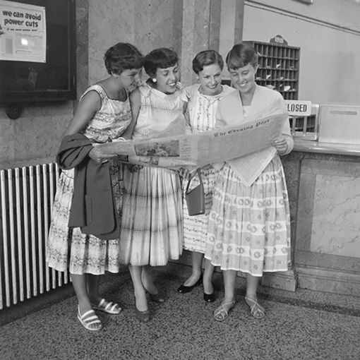 Black and white photo of four smiling young women huddled together as they read a copy of 'The Evening Post' newspaper.