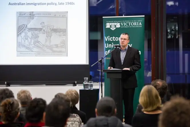 Paul Hamer speaking at the Library in front of a cartoon relating to Australian immigration policy.