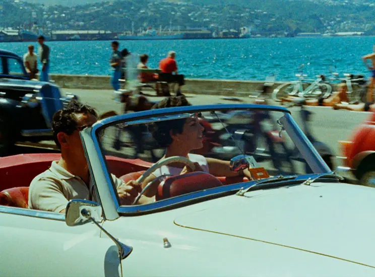 A man and woman driving in an open-top car past the ocean.
