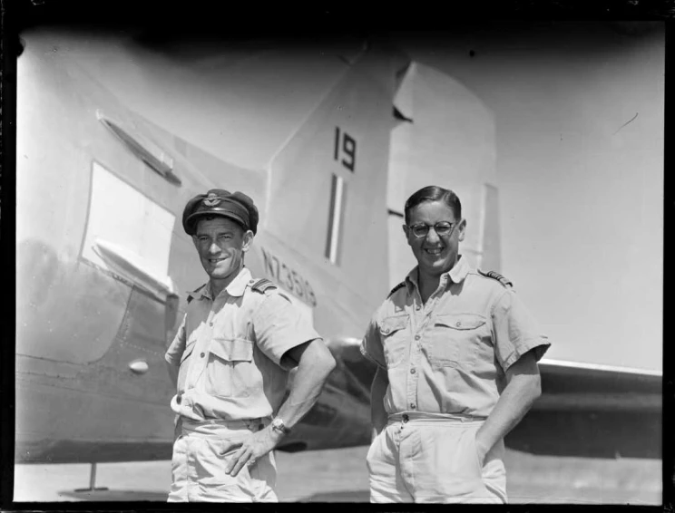 Two pilots standing side by side in short sleeved uniform with the tail section of a plane visible in the background.