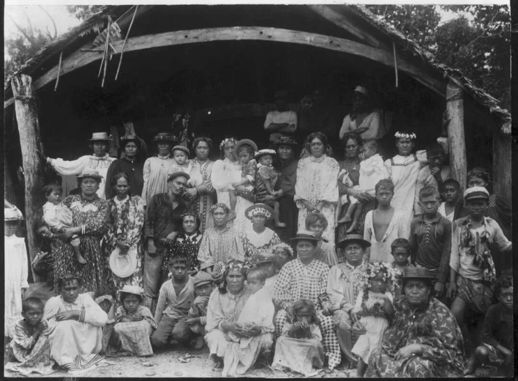 A large group portrait in black and white with people of all ages, both seated on the ground and standing.