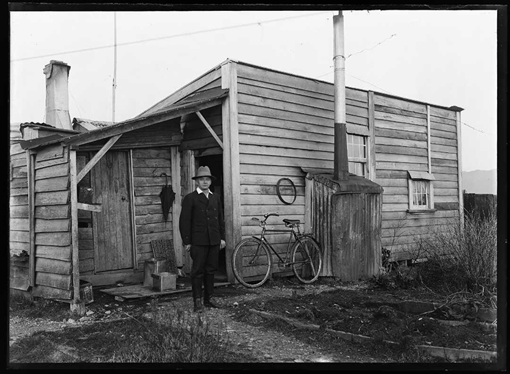 Black and white photo of a man in a suit and hat standing in front of a wooden cottage. There is a bicycle leaning against the outside wall. 
