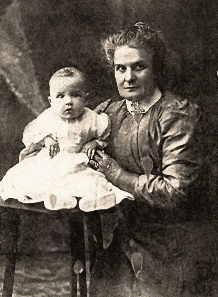 A formal black and white portrait of a woman who is crouched beside and baby, who is being supported on a stool. 
