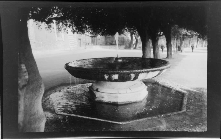 A fountain in the shape of a large, raised bowl is filled with water that is dripping over the edge into a larger pool that is octagon-shaped nearly at ground level.