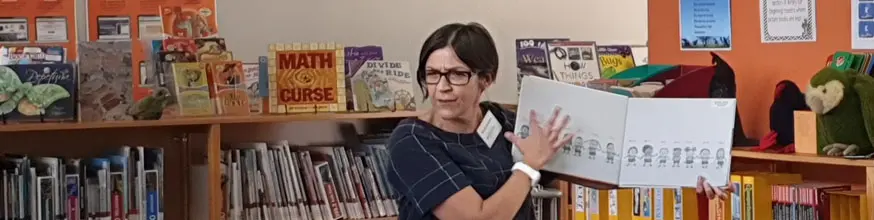 Woman reading a book aloud, she is holding a book up so the audience can see the pictures. 