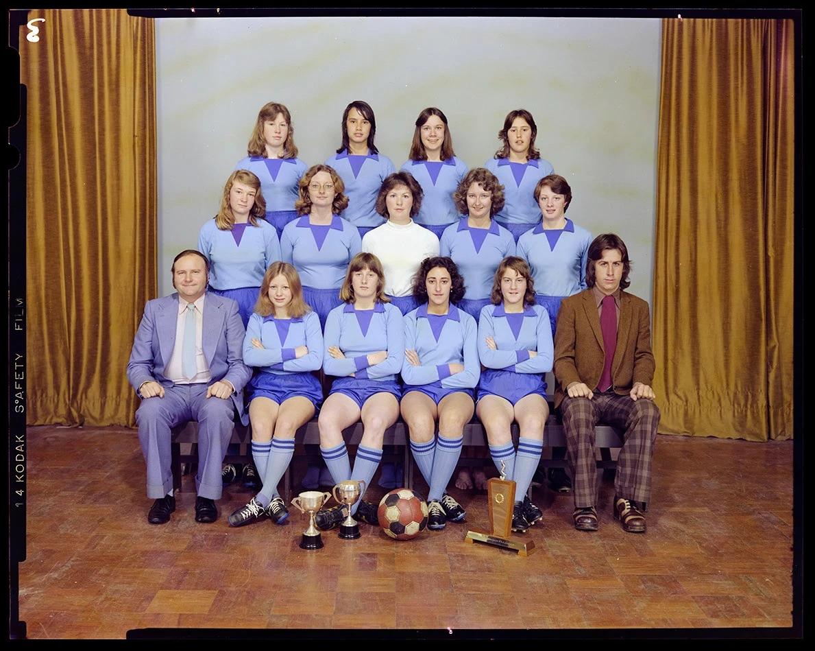 A colour studio portrait of a women's soccer team wearing blue uniforms in three rows, with the captain in middle row wearing white jersey and two male coaches seated at either end of the bottom row. 