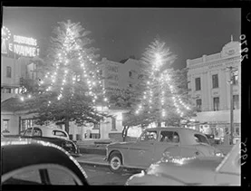 Two illuminated Christmas trees in a town square at night, with vintage cars and buildings visible in the background.