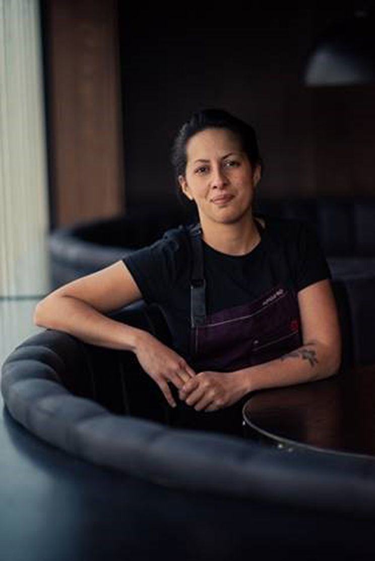 Woman sitting at a table, she is wearing a chef's apron.