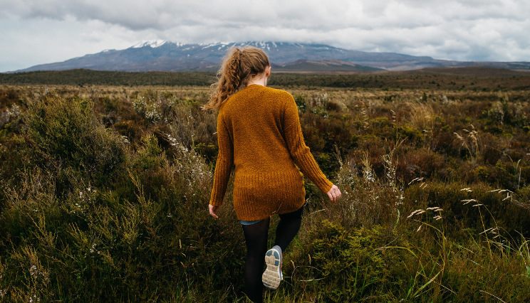Tongariro peak shrouded in clouds in the background with a girl in brown jersey stepping towards the mountain in the foreground