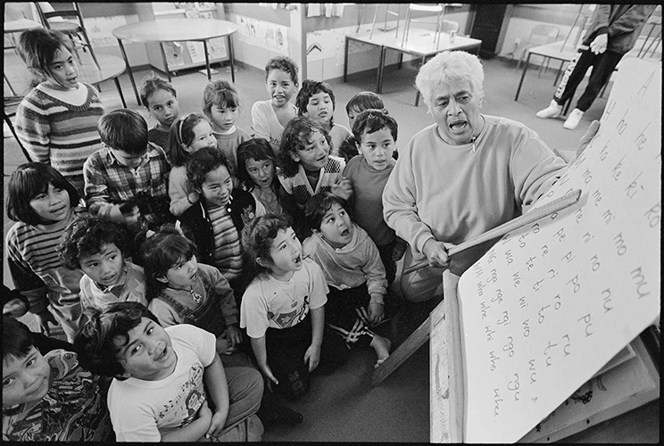 Black and white photo of female teacher pointing to a poster with words in te reo Māori to a class full of small children