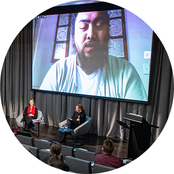 Auditorium with a Māori man appearing on Zoom on a big screen. Two women sit in chairs in front of the screen. 