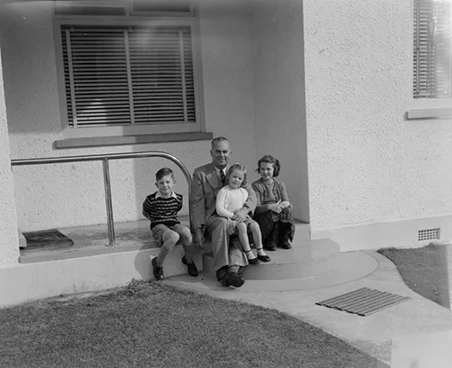 Three children and a man sit on the front steps of a stucco house.