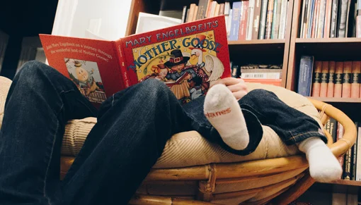 Father and child's legs and sock-covered feet sticking out from chair behind large red 'Mother Goose' book cover as they sit in a chair with bookshelves in the background