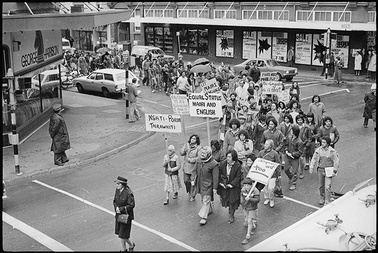Black and white image of group of protesters on 1980s clothing holding signs in support of equal status of the Māori language with English