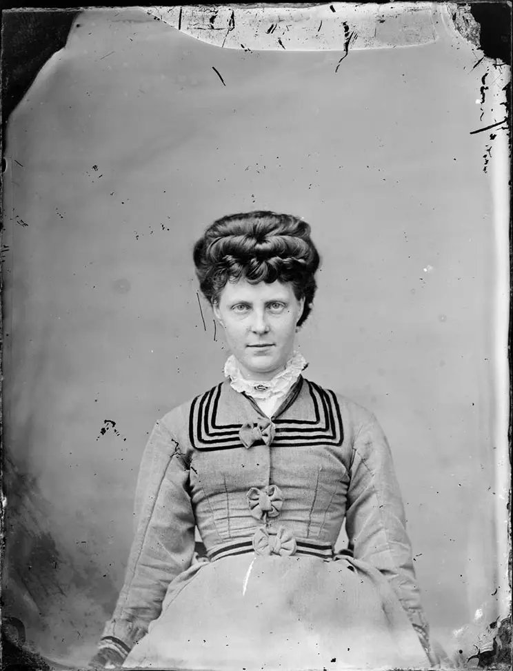 Black and white Victorian-era studio photograph of a woman wearing a jacket with a sailor boy collar, a matching skirt and a ruffled high neck blouse. She has a Mona Lisa smile.