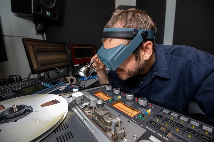 AV technician seen wearing magnifying goggles and shining a torch into the reel to reel tape machine.