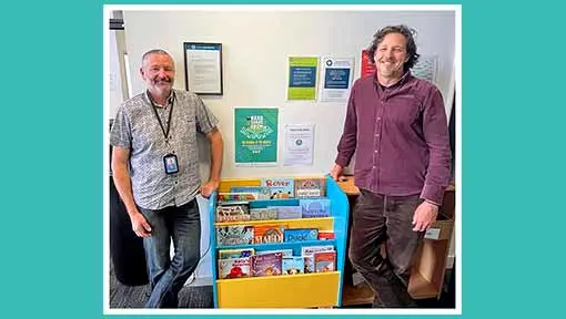 2 men smiling and standing beside a book stand with children's books.