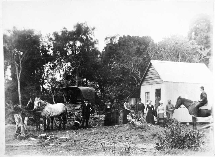 People around a carriage and horse, small wooden hut with women standing outside. Man on a horse in the foreground.