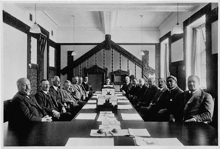 Black and white photo of men sitting at a long table in a room with Māori carvings.