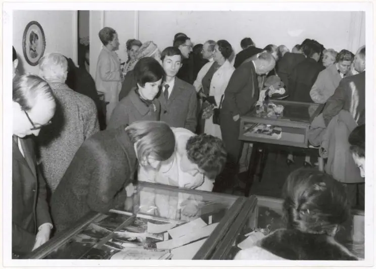 A black and white photo shows a crowded room with a few display cases and a number of the people appear to be looking down into them at the objects contained therein. 