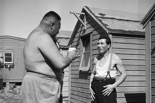 A very tall, big Māori man in shorts holding a cup of drink talking to a smaller Japanese man wearing trousers, a singlet and braces.
