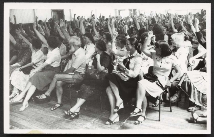 A black and white photo of a room filled with seated librarians all raising their hands affirmatively.