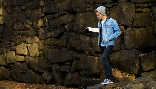 Teen boy wearing jean jacket and hat standing in front of rock wall reading book.