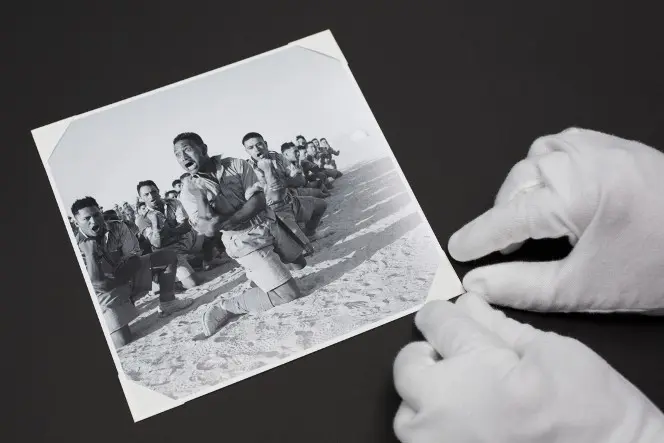 Gloved hands affixing to an album a photo of the Māori Battalion performing a haka, 1941.