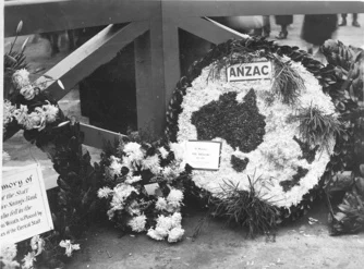 Black and white photograph of an ANZAC memorial wreath, flowers and written plaques sitting on the ground against a wooden barrier or railing]