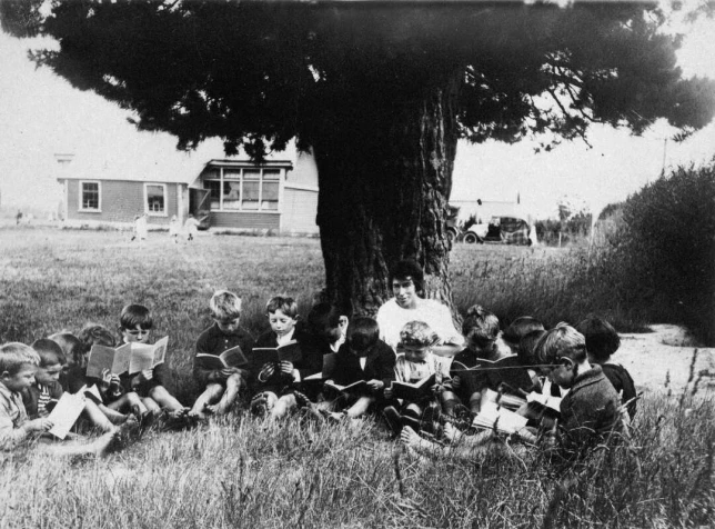 Teacher and children, reading outside under a tree, circa 1935.