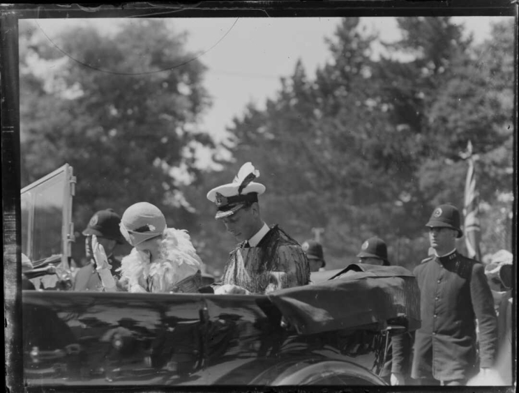 Duke and duchess are riding in the back of a car with no roof wearing a uniform and fancy dress.