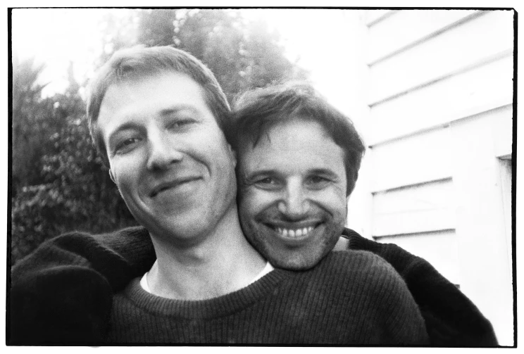 A black and white photo taken outside a weatherboard-sided house of two men hugging and smiling while facing the camera.