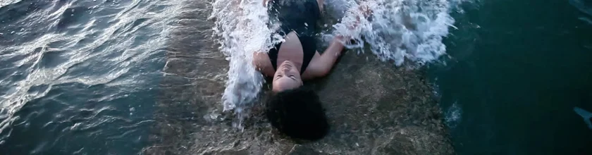 Woman lying on her back on a concrete pier. The waves are washing up on her and she is holding on trying not to get pulled into the sea. 