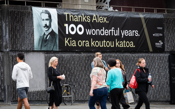 Pedestrians cross Aitken St in front of a new banner on the wall of the National Library.