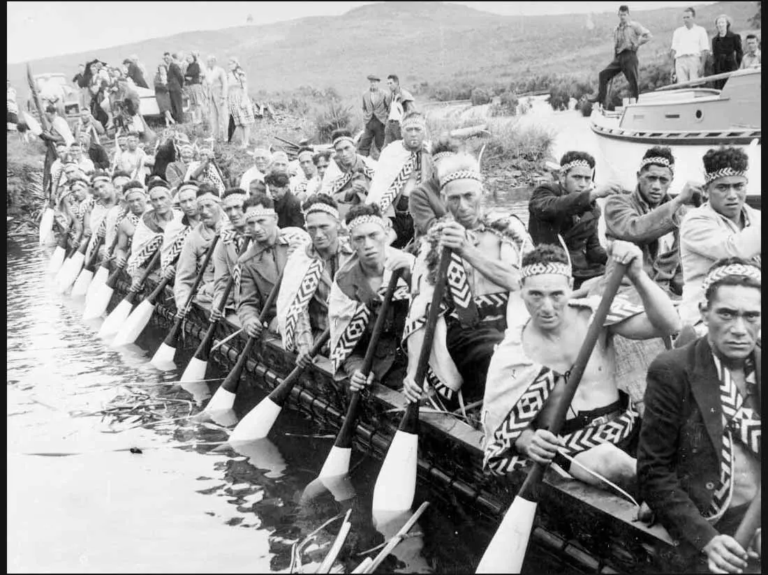 Black and white photo of a waka with Maori crew aboard, holding paddles in the water.