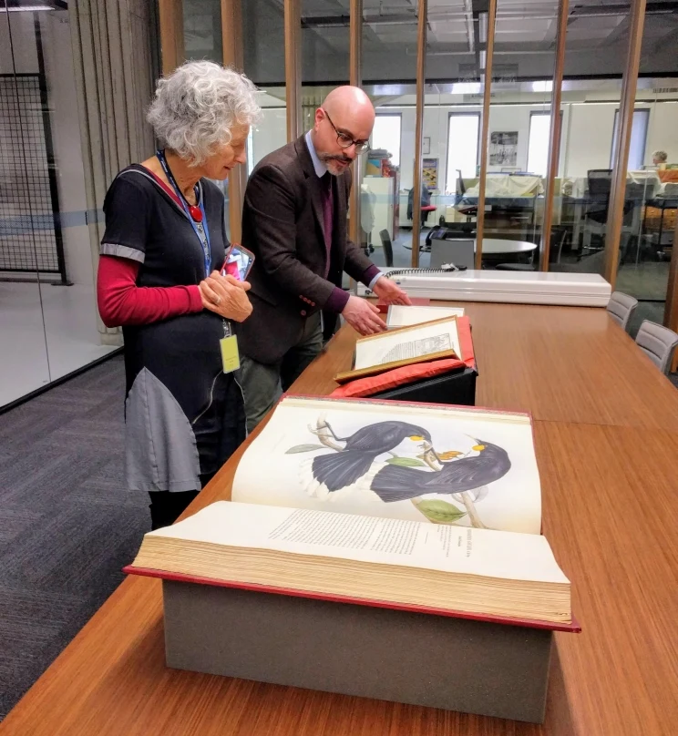 Three large bound books are displayed on a table with a curator showing them to the film director.