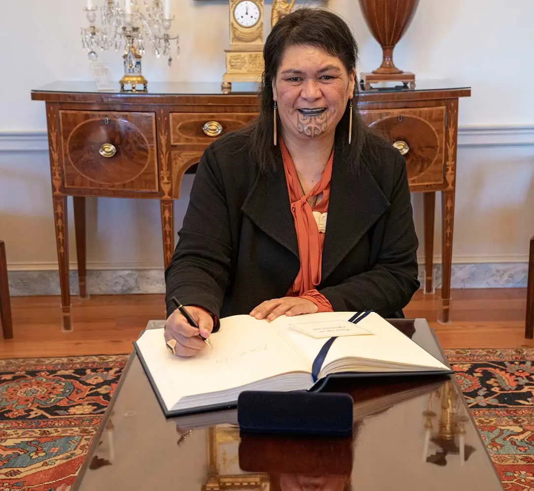 Photo of Nanaia Mahuta showing her moko kauae. Nanaia is seated, smiling and signing the US Department of State guestbook.