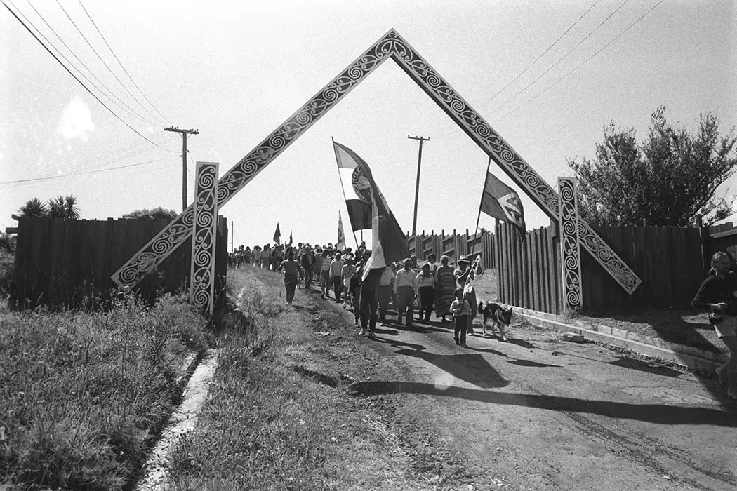 Black and white photograph of a hikoi — protestors hold flags and march from Ōrākei Marae at Takaparawhā Bastion Point.