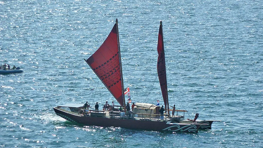 Colour photograph of the Haunui waka and its crew on the water, showing Polynesian patterns on the sails and hull.