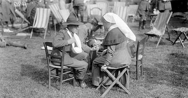 A nurse and two wounded soldiers seated in a group on wooden chairs on the grass.