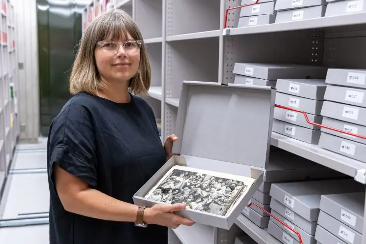 A woman stands among archival shelving stacked with grey boxes while holding open one box and displaying the black and white photographic prints contained within. 