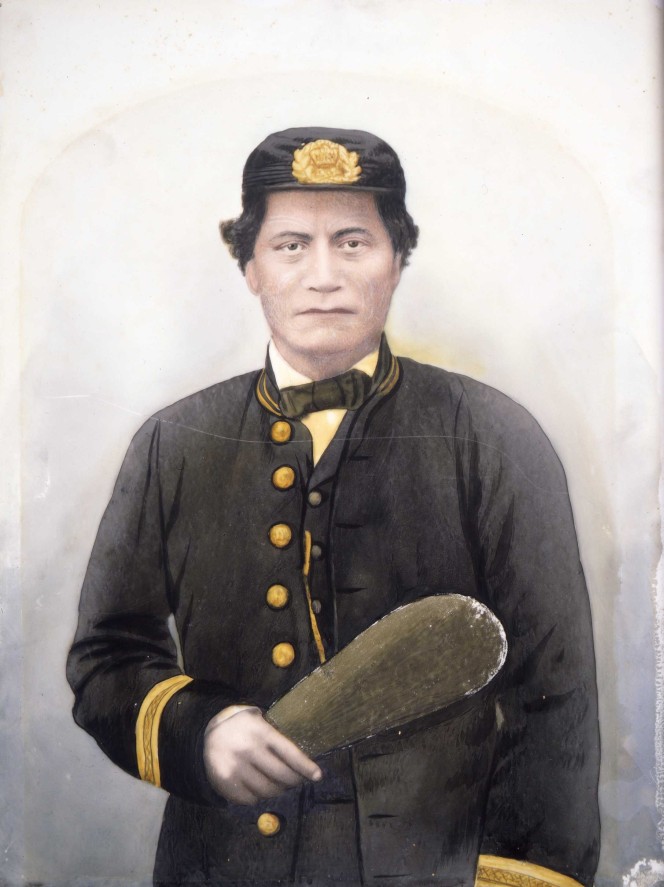 Waist-length portrait of a Maori man, identified as Rawiri Puaha, with moko, dressed in uniform, including cap, holding a mere in his right hand.