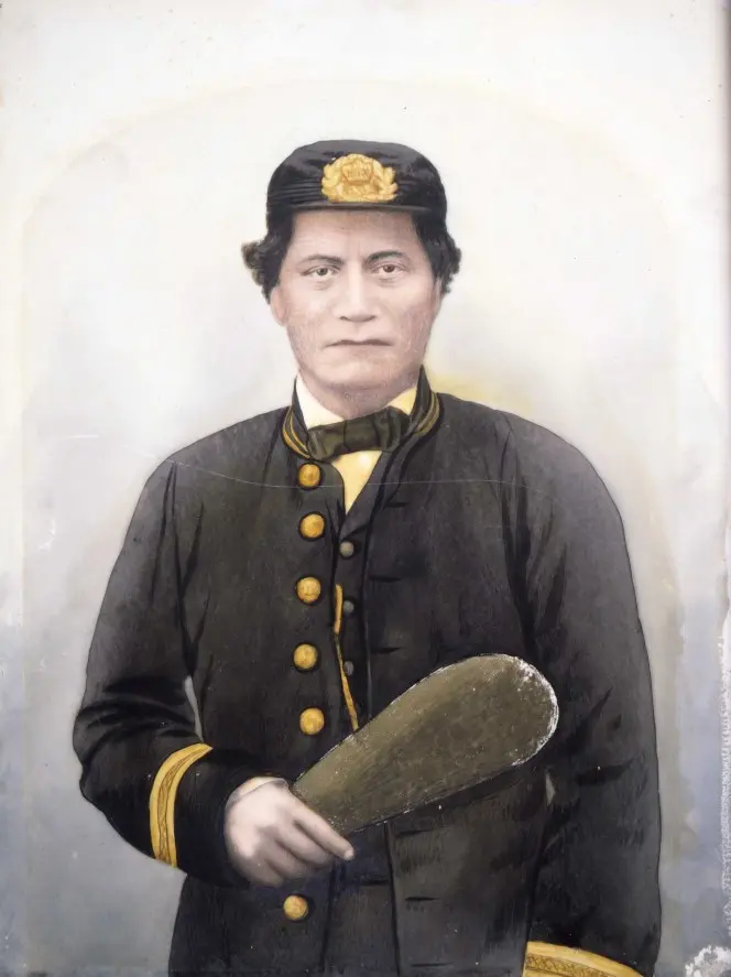 Waist-length portrait of a Māori man, identified as Rawiri Puaha, with moko, dressed in uniform, including cap, holding a mere in his right hand.