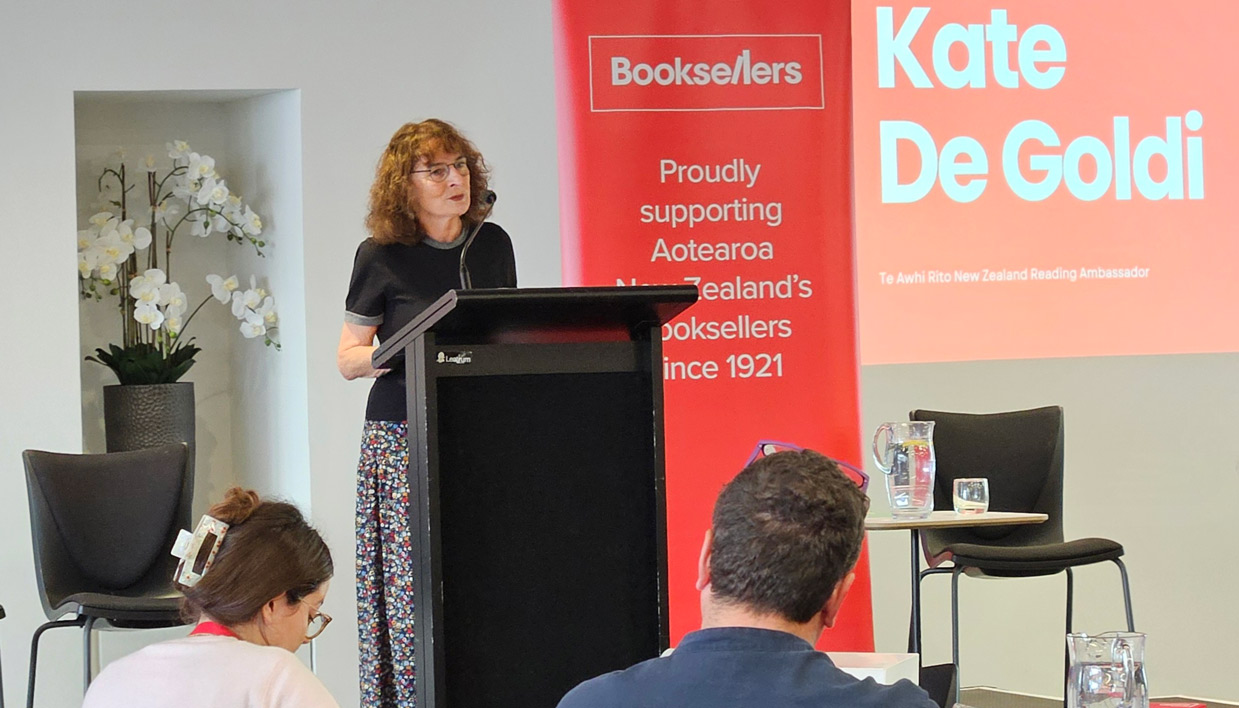 Kate De Goldi speaks at a podium at a Booksellers NZ event, with a red banner and projected slide displaying her name behind her.